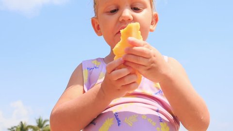 closeup little girl eats pineapple fruit standing on sand beach in resort city against azure sea