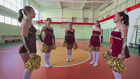 Slow motion shaky camera video, group of beautiful long legged girls jumping in circle facing inwards with pom-poms at cheerleading practice in school gymnasium, low angle