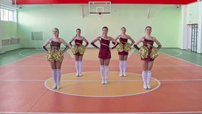 Slow-motion video of five young beautiful cheerleader girls performing in formation of circle smiling brightly and looking at camera during practice in school gym with pom-poms
