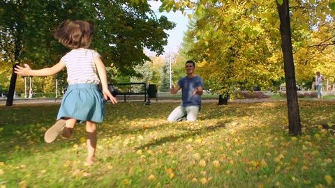 Slow motion tracking of excited little girl running on green grass and fallen leaves in park towards sitting and smiling father, then hugging him and laughing