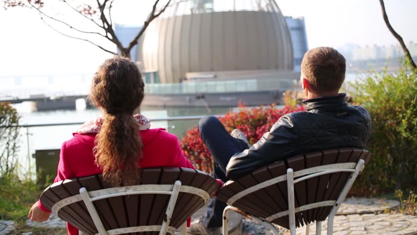 Backs of woman and man resting on chairs and looking to circle building on water in Seoul.