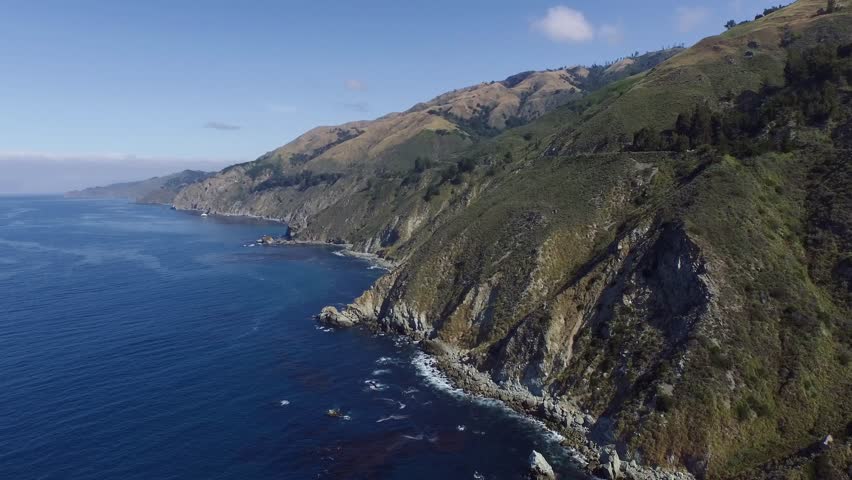 High aerial shot flying by the California Big Sur coast. Deep blue ocean and massive cliffs with waves crashing. Blue skies with sparse clouds.