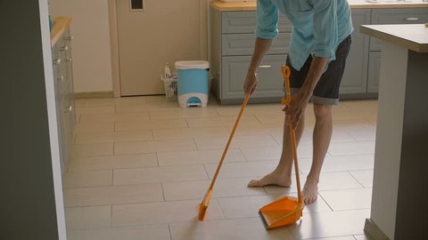 Young man, husband is sweeping the kitchen floor carefully with yellow broomstick and scoop. Slow motion, Steadicam shot