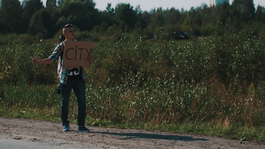 Young boy hitchhiking at road with cardboard plate city. Tourist. Thumb up. Traveler