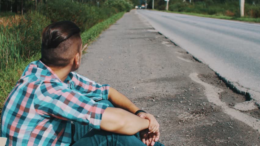 Young boy sitting at road hold ear of grass in mouth. Wait. Travel. Hitchhiking. Summer sunny day.