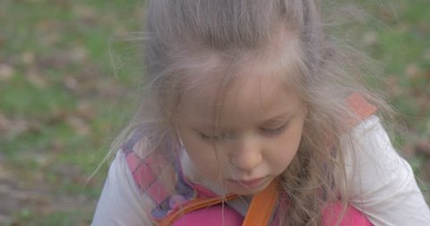 the Little Girl Sits in a Centre of a Green Lawn. She Searches an Acorns on the Ground, and Then Puts Them Into the Bag. on the Ground There Are Few Suitable Acorns.