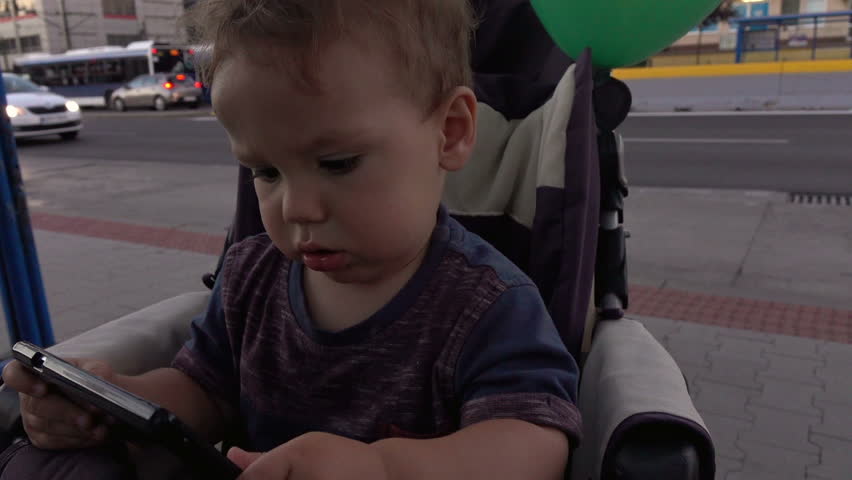 Baby is looking to smartphone, sitting in stroller at the bus stop
