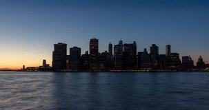 New York City Lower Manhattan skyscrapers between sunset, dusk and nightfall. Time lapse cityscape view of the Financial District lights and East River with passing boats