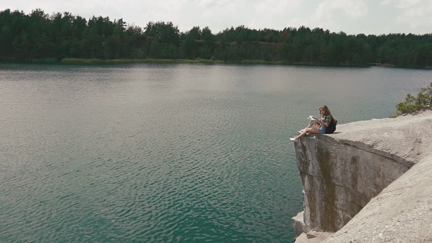 Young active backpacker girl relaxes by resting on mountain rock upon large water surface of mountain forest lake