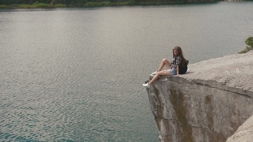 Young attractive backpacker girl relaxes by resting on mountain rock upon lake water surface below