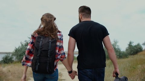 Back view of beautiful young couple with backpack walking along the road and holding hands
