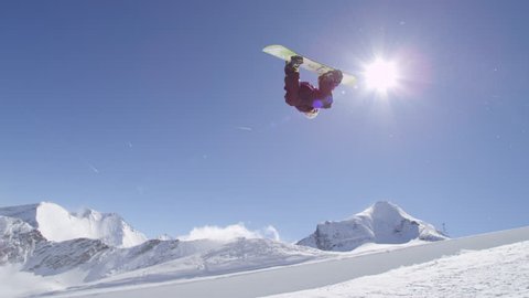 SLOW MOTION: Young pro snowboarder riding the half pipe in big mountain snow park, jumping out of the halfpipe wall and over the sun, performing tricks and rotations with grabs in sunny winter