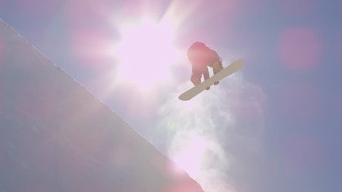 SLOW MOTION: Young pro snowboarder riding the half pipe in big mountain snow park, jumping out of the halfpipe wall and over the sun, performing tricks and rotations with grabs in sunny winter