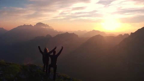 Aerial - Flyover silhouette of a couple with raised arms celebrating successful climb on the mountain in beautiful sunset