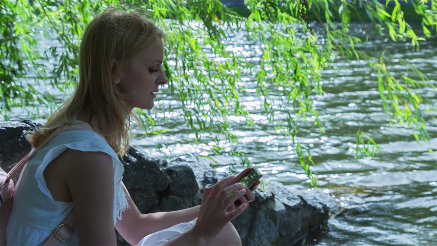 High quality 10bit footage of Beautiful girl using SmartPhone sitting near lake in city park on sunset. Made from 14bit RAW.
