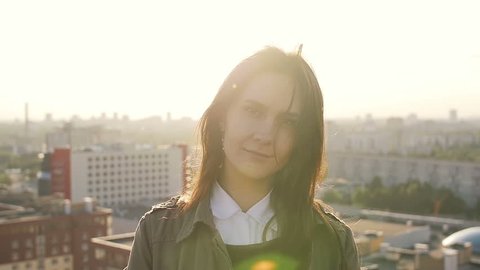 Young beautiful woman standing on the roof of high-rise building at sunset looking at the camera. Slow mo