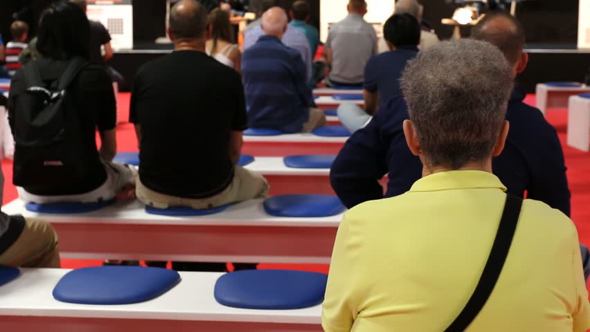 People sitting, watching and listening to host, selective focus on short hair woman in yellow t-shirt while the rest of audience are blurred