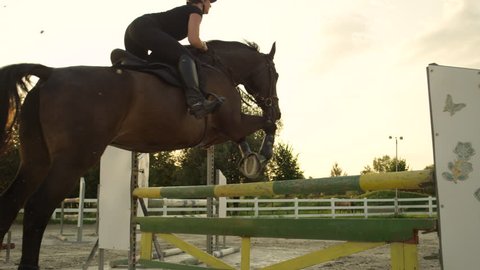 SLOW MOTION, CLOSE UP: Young horsegirl horseback riding strong brown horse jumping the fence in sunny outdoors sandy parkour riding arena. Competitive rider training jumping over obstacles at sunset