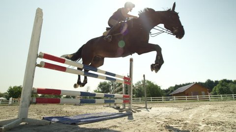 SLOW MOTION, CLOSE UP, LOW ANGLE: Horsegirl riding strong brown horse jumping the fence in sunny outdoors sandy parkour riding arena. Competitive rider training jumping over obstacles in manege