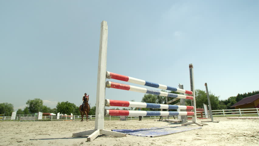 SLOW MOTION, CLOSE UP, LOW ANGLE: Horsegirl riding strong chestnut horse jumping the fence in sunny outdoors sandy parkour riding arena. Competitive rider training jumping over obstacles in manege Royalty-Free Stock Footage #19304767