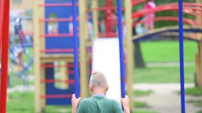 Anonymous little kid sitting on swing in public children playground on summer warm day. Blurry child playing alone outside in city urban park. Real time video footage. Shallow depth of field.