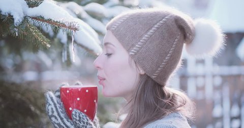 Woman Drinks Hot Tea or Coffee From a Cozy Cup on Snowy Winter Morning Outdoors. 4K DCi SLOW MOTION 120 fps. Beautiful Girl Enjoying Winter in a Garden with a Mug of Warm Drink. Christmas Holidays 