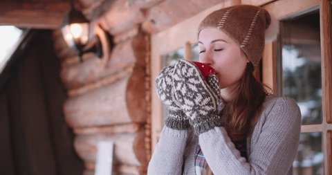 Woman with Cup of Hot Tea or Coffee Stands by the Cozy House on Snowy Winter Morning. 4K DCi SLOW MOTION 120 fps. Beautiful Girl Enjoying Winter Outdoors with a Mug of Warm Drink. Christmas Holidays 