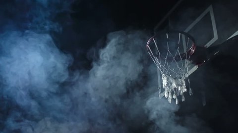 Basketball player jumping in the air and performing reverse dunk in the dark court in slow motion with smoke in the background