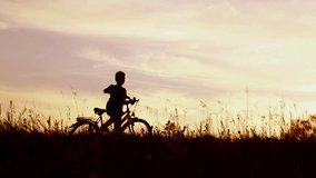 Little kid riding bike in summer meadow landscape at beautiful sunset sky background. Silhouette of anonymous caucasian small boy of 9 years old sitting on bicycle in countryside area. Real time video