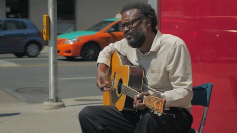 Toronto, Canada - CIRCA: August, 2016: Close up of black male with a beard busking by sitting and playing a guitar on a sidewalk in front of a bank while pedestrians walk by