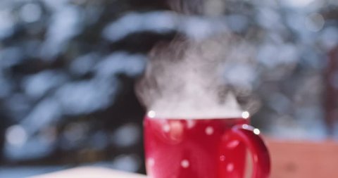 Steaming Cup of Hot Coffee or Tea standing on the Outdoor Table in Snowy Winter Morning. 4K DCi SLOW MOTION 120 fps. Cozy Festive Red Mug with a Warm Drink in Winter Garden. Christmas Morning Concept