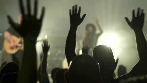 Stock video footage Crowd Raising hands at a concert, focus is on the hands, band is out of focus in background. silhouetted people.