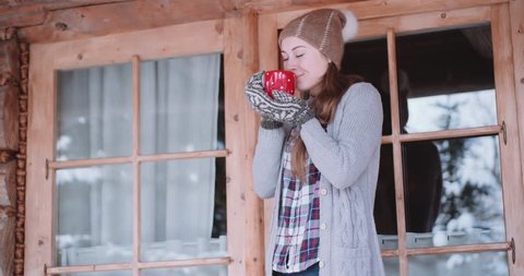 Woman with Cup of Hot Tea or Coffee Stands by the Cozy House on Snowy Winter Morning. 4K DCi SLOW MOTION 120 fps. Beautiful Girl Enjoying Winter Outdoors with a Mug of Warm Drink. Christmas Holidays 