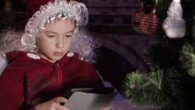 Christmas night, a little boy with a santa hat lying on a cushion plays on a tablet, at background the christmas tree illuminated with christmas gifts on the floor, the atmosphere is warm and cozy