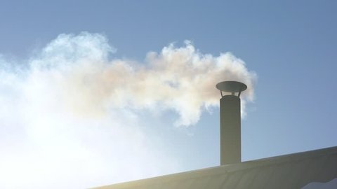 Smoke comes from the chimney of the house. The pipe on the roof. Chimney. Country house. The house with a chimney. Smoke in the blue sky. Smoke stacks and sky clouds