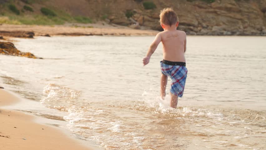 A slow motion shot of a little boy running across the ocean beach through the water