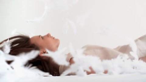 Young woman falling down on the bed with white flying feathers 