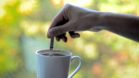 Woman hand mixing of hot coffee with a spoon in a mug near the window glass. Blur autumn background