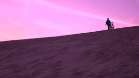 Man with his bike walking over sand dune, purple sunset colors. 4K video