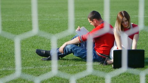 young man reading a book - a pretty girl working on a laptop on the football field