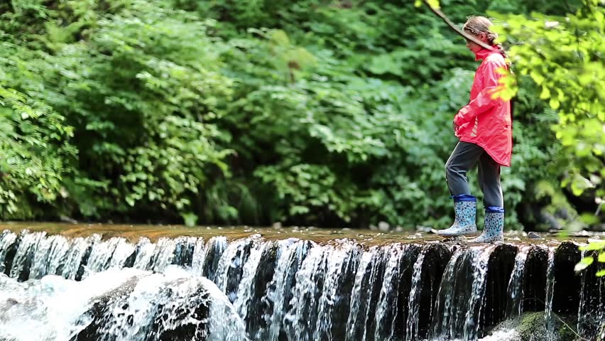 Girl in red jacket walks along small overflow dam on the river in the forest. Girl walks on the brook in the wood