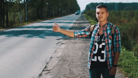 Young man with backpack hitchhiking at road in countryside. Thumb up. Sunny day. Problem