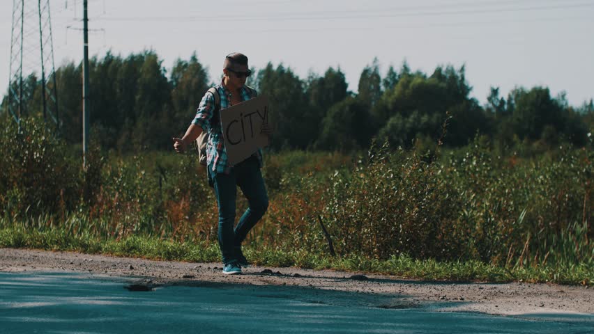 Man walk along the road with cardboard plate sign city. Hitchhiking. Thumb up. Cars. Travel. Trouble