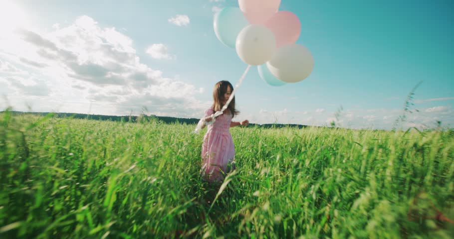 Little Asian girl in a dress running through green wheat field with balloons in hand, slow motion