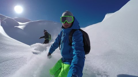 SELFIE: Cheerful snowboarders having fun snowboarding backcountry on sunny winter day in snowy mountains. Extreme freeride snowboarders riding fresh powder snow and doing powder turns off piste
