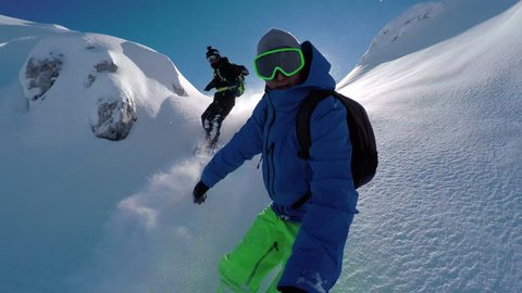 SELFIE: Cheerful snowboarders having fun snowboarding backcountry on sunny winter day in snowy mountains. Extreme freeride snowboarders riding fresh powder snow and doing powder turns off piste