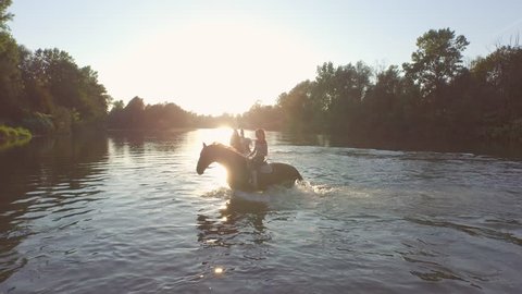 AERIAL, CLOSE UP: Three cheerful, smiling girls riding palomino and dark brown horses along green overgrown river bank and having fun on summer vacation. Happy girlfriends on horses playing in water