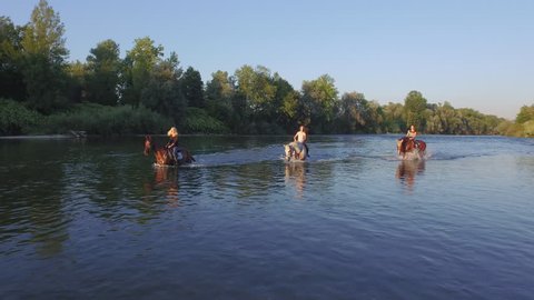 AERIAL, CLOSE UP: Three cheerful, smiling girls riding white and dark brown horses along green overgrown river bank and having fun on summer vacation. Happy girlfriends on horses playing in water