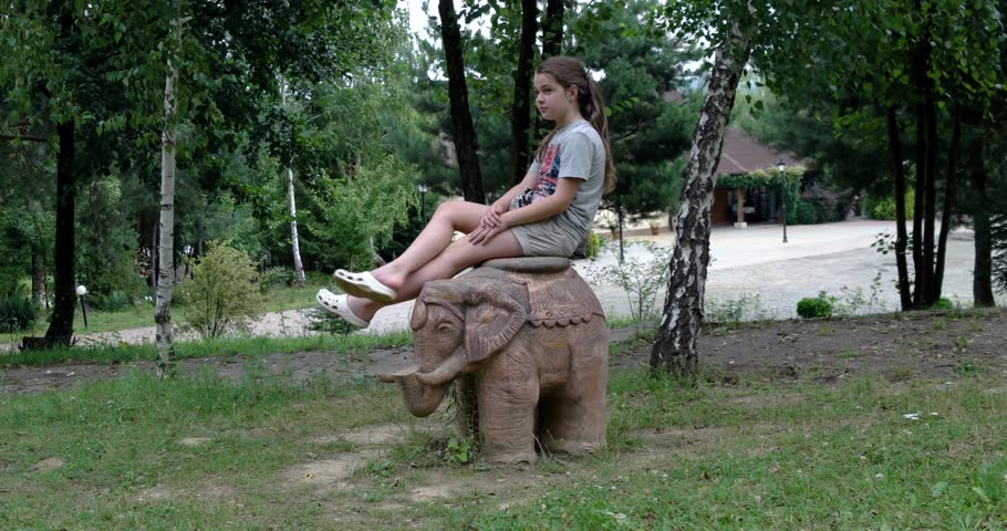 Little Brunette Girl Sits and Pose For the Camera on on Stone Elephant Sculpture in Park