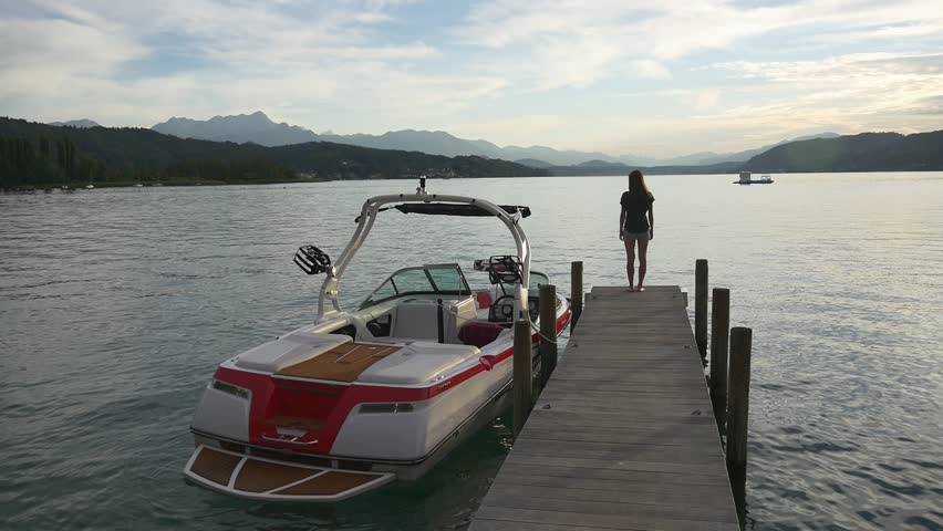 Pensive woman stands near motorboat moored to jetty looking into the distance to picturesque landscape of glaring water and mountains